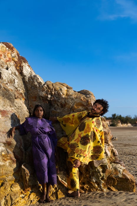 A woman in a purple dress and a man wearing a yellow patterned outfit stand in front of a rock