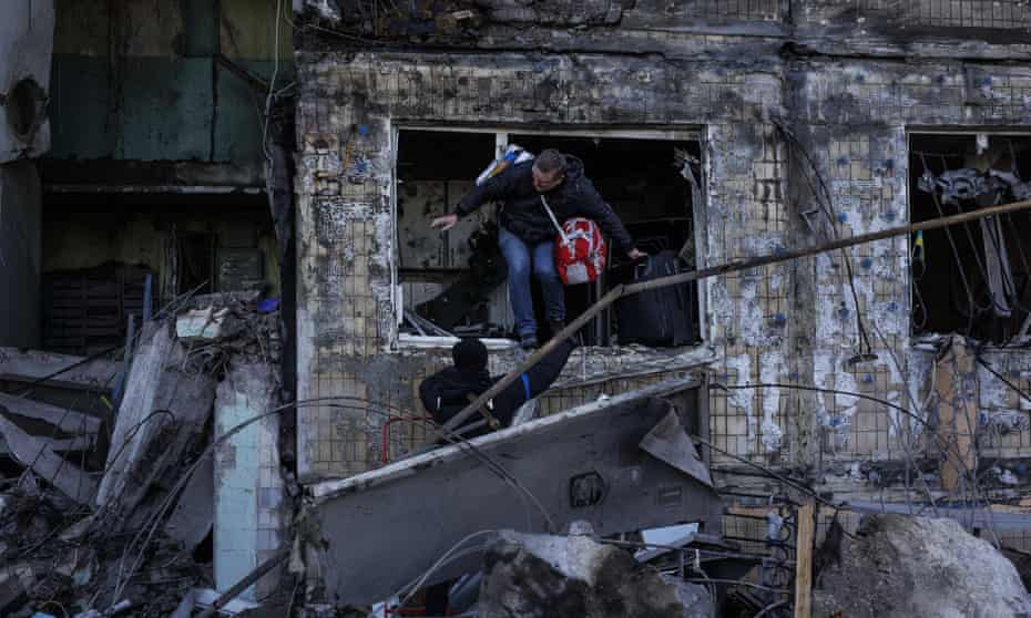 Man with wheelie suitcase climbing out of destroyed residential building.