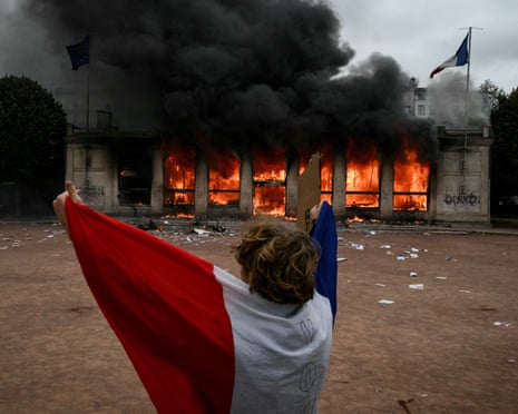 A protester wrapped in a French flag faces Lyon's burning tourism office during a demonstration