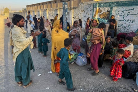Men, women and children wait by a high wall; they have bags of possessions and wear coloured long robes and shawls. One young girl wears a red embroidered dress and some boys wear dark green long shirts and trousers.