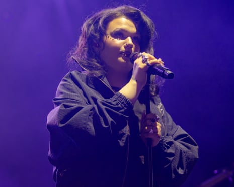 Lola Young in a dark jacket sings into a microphone against a purple-lit background