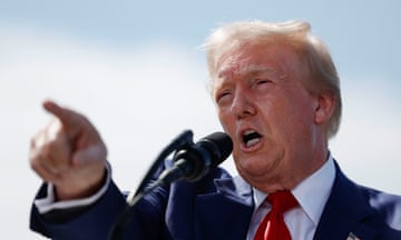 Donald Trump, a man wearing a blue suit and red tie, speaks into a microphone and points ahead