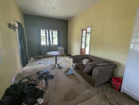 interior of a house with rubbish and dirt strewn over the floor, a broken sofa and a table holding stacks of videotapes