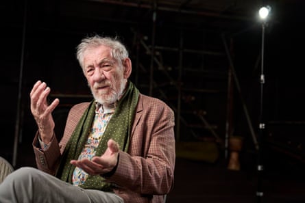 Ian McKellen gesticulates with his hands as he speaks and looks animated. He is sitting in a dark space with a spotlight behind him. He has a grey beard and wears a patterned shirt, brown checked jacket and a green silky scarf with white polka dots.