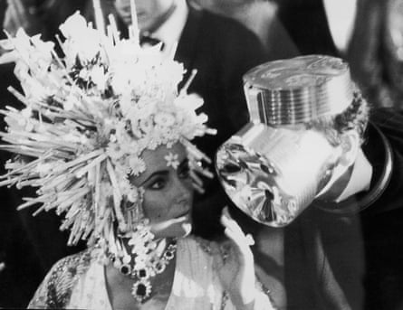 Elizabeth Taylor in a headdress of silver spikes, orchids and lilies chats to designer Cardin, who is wearing what looks like an aluminium gas mask.