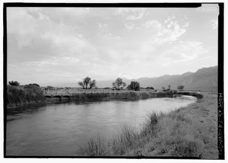 black and white image of river with low plants on bank and mountains in the background. a bridge or walkway runs along it