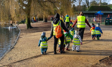 A group of children from a nursery school in high visibility jackets walking with teachers or childcare workers through a park in winter