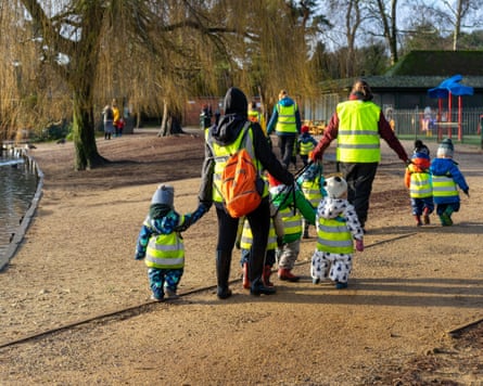 A group of children from a nursery school in high visibility jackets walking with teachers or childcare workers through a park in winter
