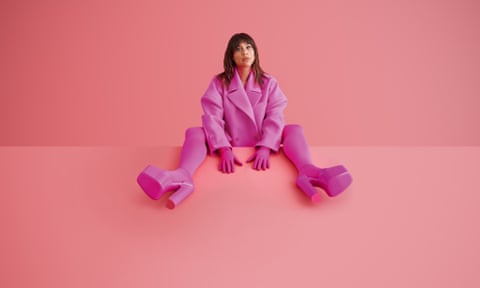 Zawe Ashton wearing a shocking pink jacket, gloves, tights and shoes, with her legs up on the table in front of her, shot against a pink backdrop