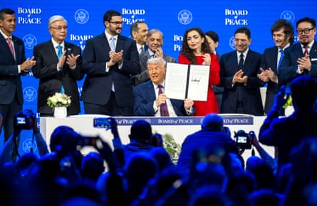 A seated Donald Trump holds up a signed founding charter as men, and one woman, stand behind applauding