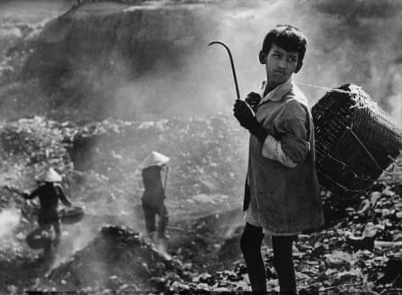 A boy stands amid piles of smoking garbage with a basket slung over his shoulder and a hook in his hand