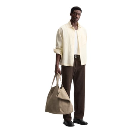 A model wears a butter yellow open shirt, with a low neck white T-shirt. A pair of dark brown trousers and a taupe holdall.