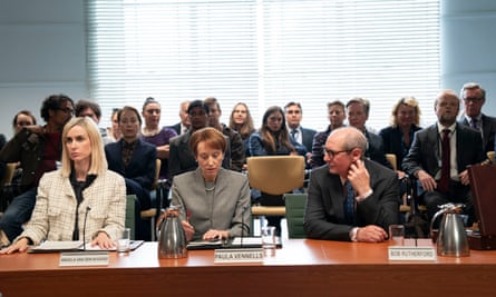 Courtroom scene from Mr Bates vs the Post Office with Katherine Kelly in a suit jacket sitting at a long desk with microphones next to Lia Williams as Paula Vennells with pearl earrings and Ian Hart, wearing glasses, in front of a crowd of seated people with venetian blinds behind them