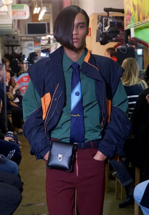 A male model on the catwalk in a green shirt, blue tie, blue and orange bomber jacket and burgundy trousers