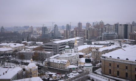 wide view over Kyiv from the Great Lavra Bell Tower, showing a church, streets and blocks of historic buildings, with a landscape of more modern tall towers behind against the skyline, under a light layer of snow and grey, wintry light
