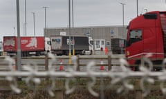 Trucks parked in a large car park in front of a building, with a high security fence out of focus in the foreground