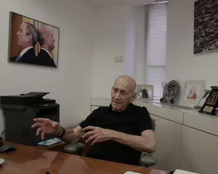 he sits behind a long wooden table or desk with white cupboards, a printer, and photographs behind him. He is nearly 80, bald, and wears a black polo shirt; he is raising his hands as he speaks.