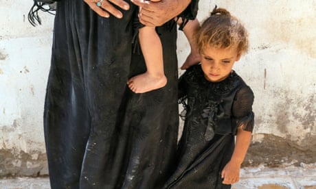 A woman holds a baby, while her daughter clings to her leg.