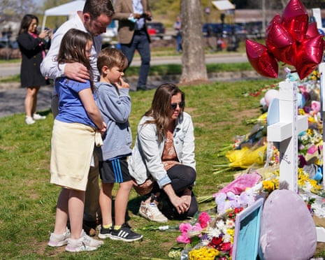 Parents and two children lay flowers by a white cross outdoors, with balloons and a squishmallow