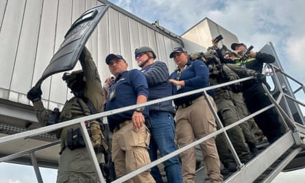 A line of at least seven police and military people walk down an outdoor stairway chest to chest, with the person in front holding a bulletproof shield up over is head, as if to protect Mancuso from being shot.