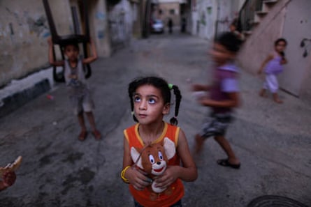 A Palestinian girl holds her toy while playing with other children in an alley