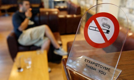 A man smokes a cigarette behind a smoking ban sign in Athens
