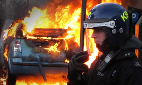 A police officer in riot gear stands near a burning car in Hackney, London
