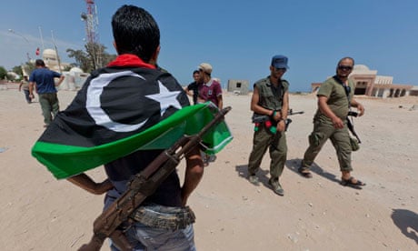 Rebel fighter in jeans with gun and new Libyan flag hung over shoulders watches his comrades