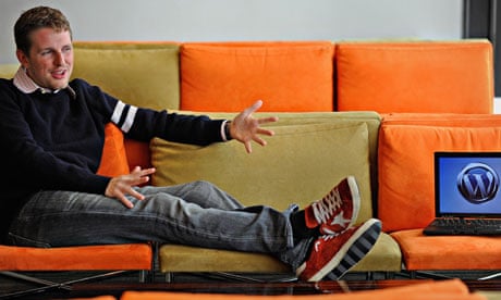 WordPress founder Matt Mullenwegg on an orange sofa pointing at a laptop with WordPress on screen