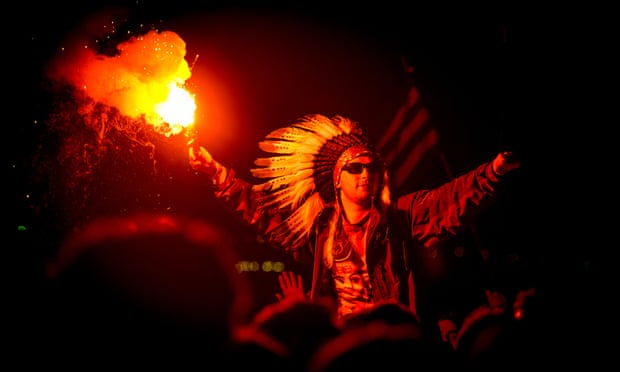 A man in the crowd wearing a Native American Headdress holding a lit flare as Jake Bug performs headlining The Other stage at Glastonbury