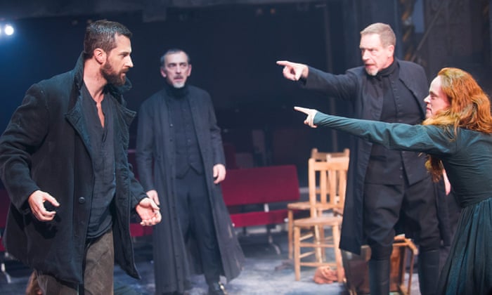 Four people are standing on a stage, three men in black clothing and one girl in a teal dress. One of the men and the woman are pointing at another one of the men.