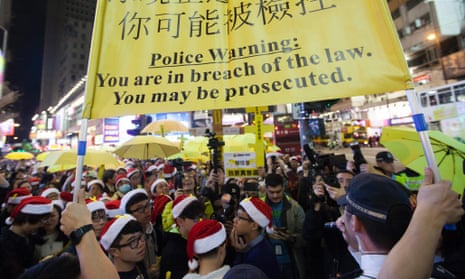 Hong Kong police hold up a warning banner as pro-democracy protesters fill a street and sing Christmas songs.