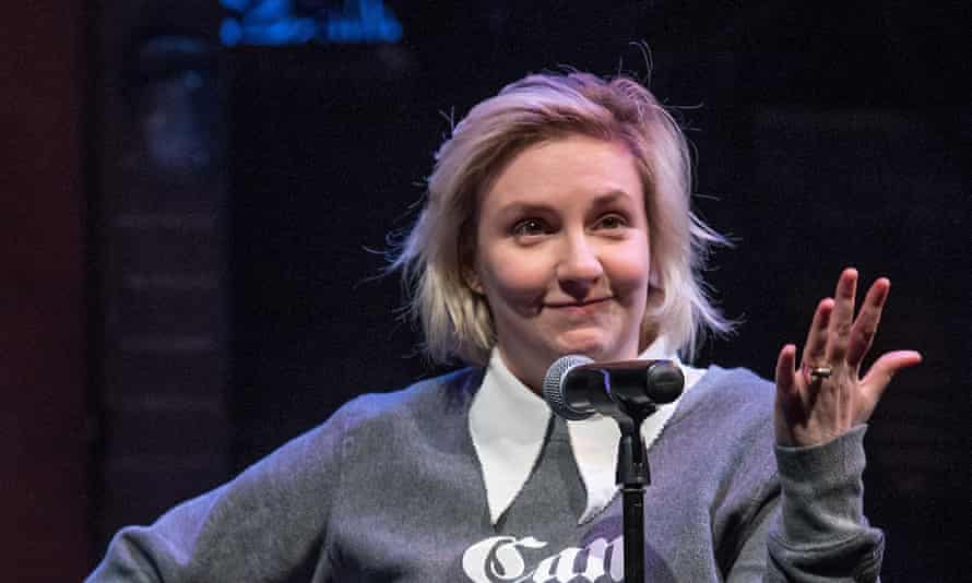 Lena Dunham in sweatshirt with smirk on her face and hand raised