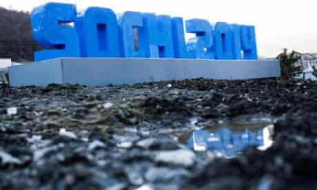 A Sochi 2014 logo standing on mud and rubble in front of snow-covered mountains is reflected in a puddle