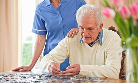 Home caregiver standing behind senior man assembling jigsaw puzzle on table