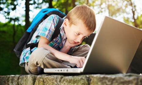 Boy with rucksack on back sits on wall in the sunshine working at a laptop