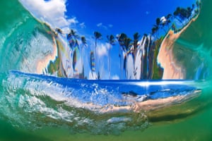 Shot from behind a breaking wave looking towards shore from an underwater perspective. The curve of the wave distorts the beach scene elongating the palm trees, in Oahu, Hawaii.