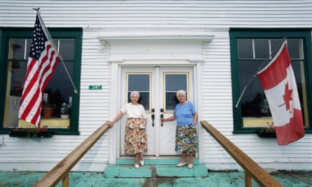 Norton, Vermont, USA --- Norton town clerks Sisters Miriam Nelson (L) and Ruth Nelson (R) stand in front of their store and office with the United States and Canadian flags. The Vermont town store is divided by the United States and Canadian border. | Location: Border of Norton, Vermont, USA and Stanhope, Canada.  --- Image by  Bob Krist/Corbis adult American American flag Canada canadian flag commerce Essex County female flag full-length general store government government official leader local government official national flag New England North America North American north american flag Northeast Kingdom Norton office Orleans County people political leader portrait Quebec room senior adult siblings sister store two two people USA Vermont women