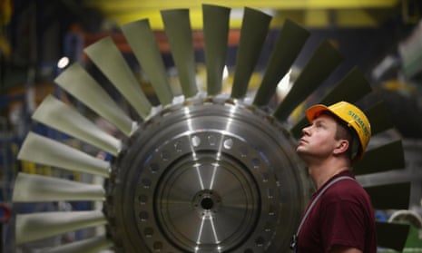 A worker watches as a part for a turbine moves above him while standing in front of a rotor assembly at the Siemens gas turbine factory in Berlin