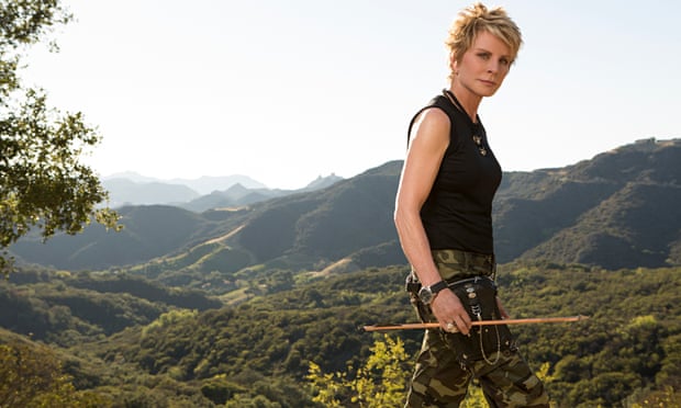 Patricia Cornwell in camoflage trousers walking, carrying an arrow, mountains behind her