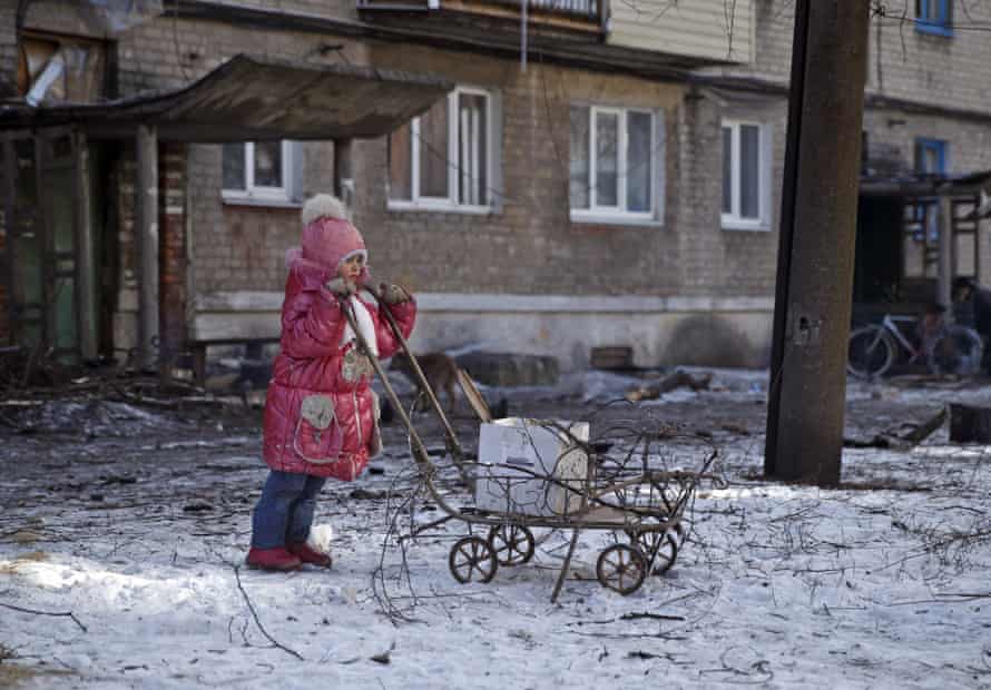 A girl leans on a cart used to carry tree branches for fire, outside a damaged apartment building