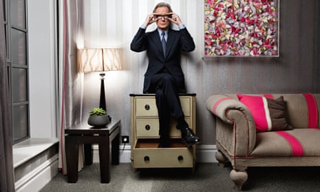 Bill Nighy holdiing his glasses, sitting on a chest of drawers with his feet in a drawer