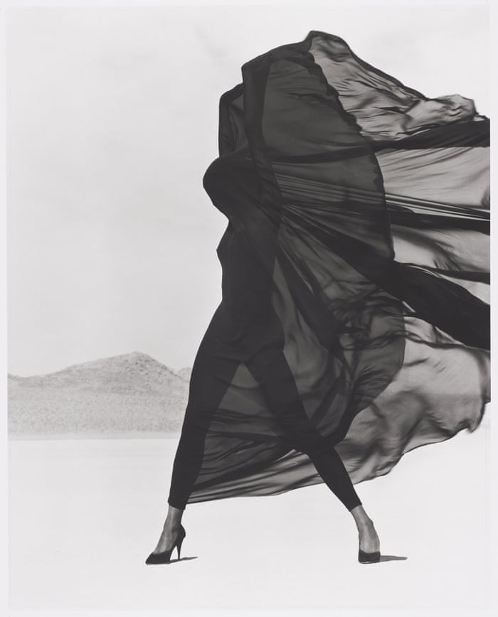 a model covered in sheer, dark fabric billowing in the wind, standing alone in a very bright desert