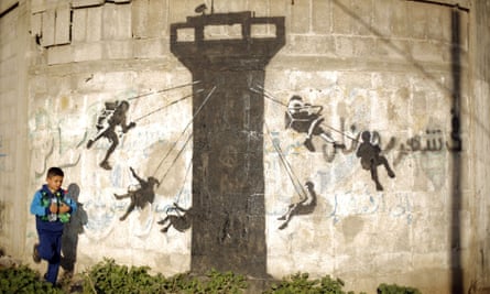 A Palestinian child walks past a mural of children using an Israeli army watch tower as a swing ride, said to have been painted by Banksy.