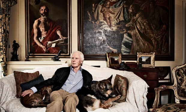 Brian Sewell on a sofa with his dogs at home, paintings behind
