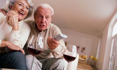 Two old people watching television with glasses of wine. 