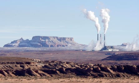 Bare rock and earth in foreground, red/brown, rock outcrops at the horizon, and to the right three tall chimneys and industrial buildings billowing smoke.