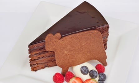 Slice of rich layered chocolate cake next to a cow-shaped biscuit, some cream and raspberries, blueberries, strawberries