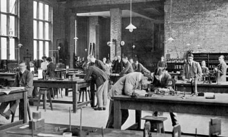 Black and white photograph of the inside of a large, high-ceilinged brick building.  Lights hang down from long chains over wooden tables where students – mostly male – in Edwardian suits and coats, examine technical equipment.  Two possibly female figures can also be seen.