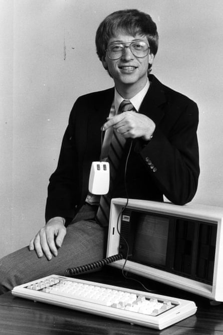 A young and smiling Bill Gates perched on a desk with a computer on it